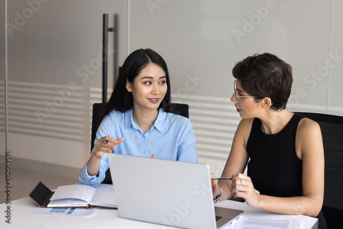 Two positive diverse female business colleagues talking at laptop, using modern technology for teamwork, discussing online startup project, cooperation strategy, sitting at meeting table