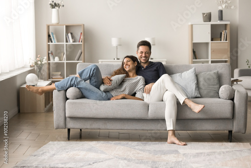 Positive happy young husband and wife relaxing on comfortable couch together, talking, hugging, laughing, having fun, enjoying romantic leisure, relaxation at home, good warm relationships