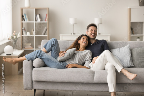 Positive happy young husband and wife relaxing on new comfortable pale grey sofa, hugging, talking, laughing, having fun, spending home funny leisure time together