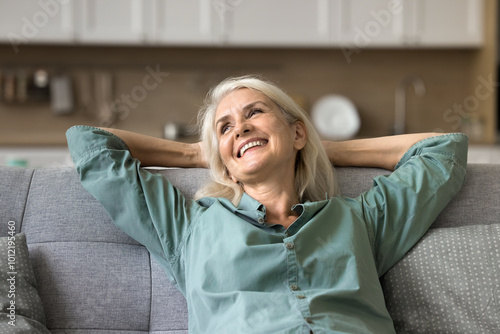 Cheerful aged woman resting on cozy couch with smile on her face, looking away feel happy, spending untroubled time at home, enjoy comfort and smart home climate control and silent domestic appliances