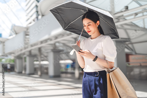 Business asian woman holding UV protection umbrella with shopping bags using smartphone on sunny day