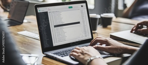 Close-up of a person's hands typing on a laptop computer with an email inbox open on the screen, while sitting at a wooden table.