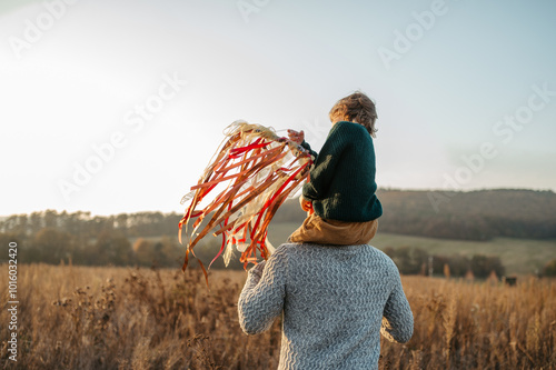 Rear view of father walking with daughter on his shoulders across autumn meadow. Girl holding wreath with ribbons.