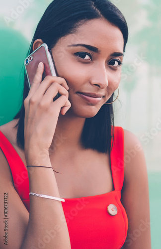 Close up portrait if attractive Latin hipster girl looking at camera while making international mobile conversation, young millennial phoning via contact application while posing near publicity area