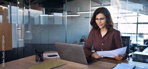 Focused latin hispanic business woman working on laptop computer reading financial document report in office. Accountant entrepreneur manager businesswoman doing paperwork using pc. Copy space, banner