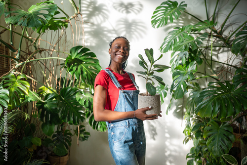 Smiling African American woman holds potted ficus, surrounded by lush flowers and plants in home garden. Black female florist caring of plants, Indoor greenery, hobby, nature lover, mindful living