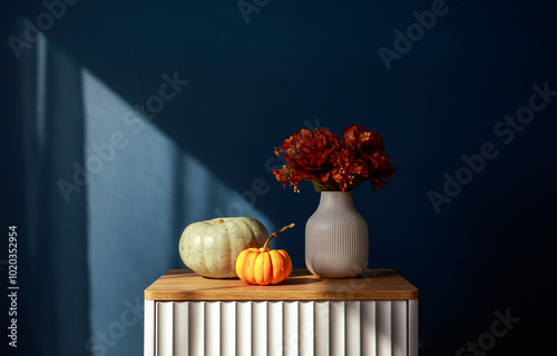 Autumn still life with green and orange pumpkins, vibrant flowers in a white vase, and dramatic shadows on a blue wall. Perfect for seasonal decor and harvest themes