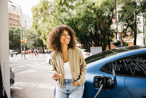 A woman is smiling as she stands next to her electric car at a charging station in a city environment. The scene captures modern transportation and eco-friendly living.