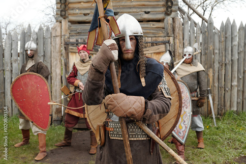 Group of medieval armored warriors standing in front of wooden fort entrance, holding various shields and weapons while wearing helmets and chainmail ensemble