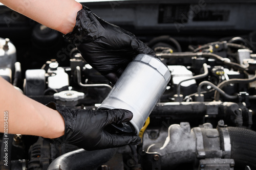 Auto mechanic repairman in working gloves holds fuel filter for diesel cars against background of car engine compartment, close-up. Auto parts replacement concept, car maintenance, repair services