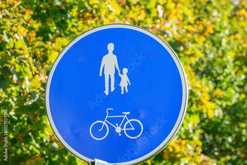 A blue circular sign indicates an area designated for pedestrians and cyclists, highlighting safety for families walking together in a park setting