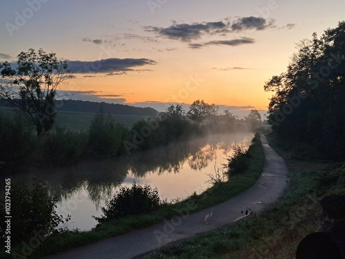 Magnifique paysage de campagne au crépuscule : un chemin sinueux longe un canal paisible, baigné dans une douce lumière orangée. La brume matinale ajoute une touche de mystère à la scène