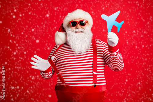 Photo of old man grey beard demonstrate blue paper plane funny wear santa claus x-mas costume suspenders sunglass striped shirt cap isolated red color background