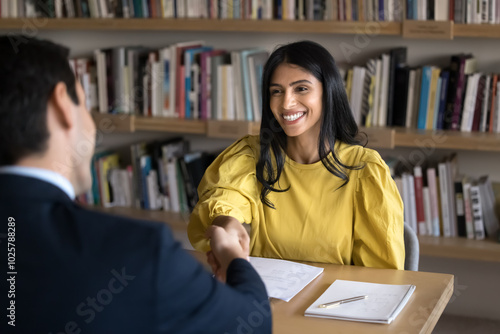 Cheerful Indian student woman talking to male professor in college library, giving handshake to tutor, teacher, thanking for help in study research, sitting at table with bookshelves behind