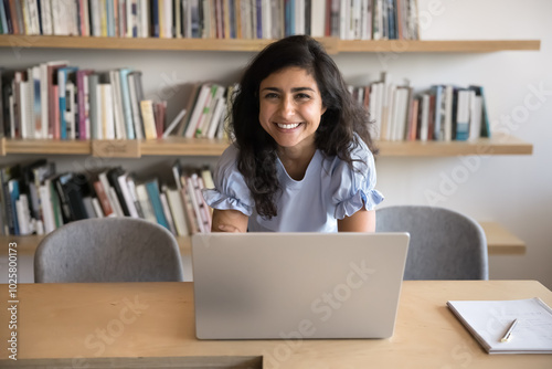 Happy beautiful Indian student woman working at laptop in library, posing for portrait at workplace table, looking at camera, bending over desktop, smiling, laughing