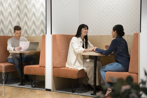 Diverse entrepreneurs, freelancers working in shared office, sitting at workplaces in co-working separate booth, meeting, talking, discussing project, reviewing documents at laptop