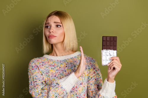 Young woman in colorful knitwear refusing chocolate bar against khaki background for shopping or dieting concept