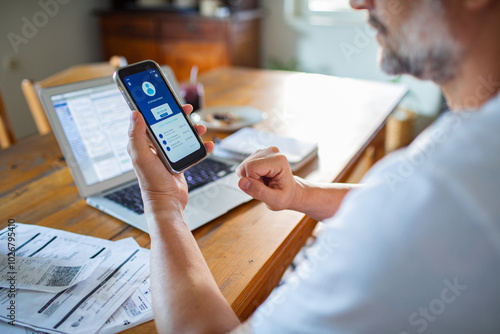 Man checking online bank account balance on smartphone while working on laptop at home