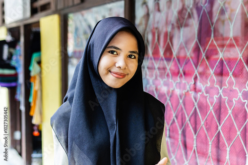 Portrait of young Asian cute muslim woman wearing black hijab smiling at camera. Joyful gen z indonesian girl standing outdoors. Indonesian ethnicity people concept