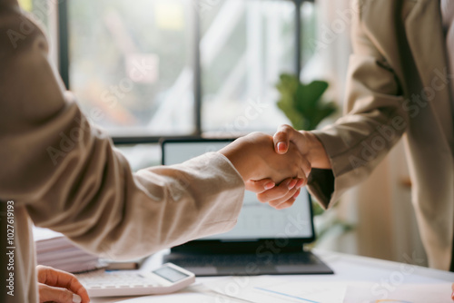 Two businesswomen shaking hands after closing a deal in their office with a laptop and calculator on the desk