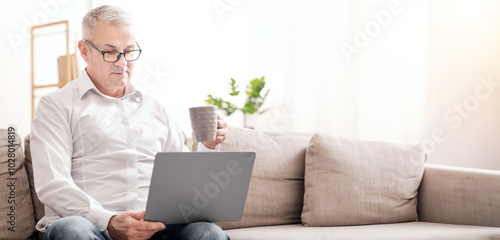 Reading Morning News. Focused Mature Man In Specs Using Laptop And Drinking Hot Tea, Sitting On Couch, Copyspace