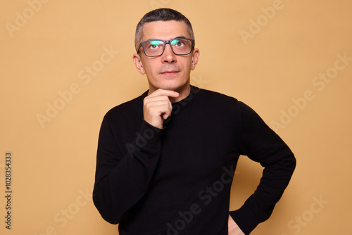 Thoughtful man in glasses and black raglan looking pensive while pondering serious idea showing quizzical expression brainstorming solution feeling uncertain and confused isolated on beige background