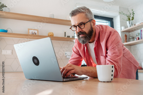 Mature man working from home on laptop, standing by kitchen island, with cup of coffee. Concept of remote work from home. Hygge at work.