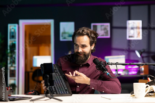 Smiling content creator filming keyboard review in personal studio using professional microphone. Happy technology specialist showcasing computer peripheral to audience on niche online channel