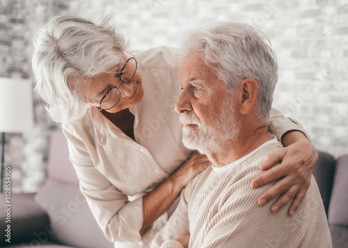 Senior woman comforting her depressed sad husband, unhappy elderly man sitting on sofa at home needed support and care. Two pensioners express their feelings and closeness