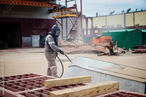 A worker sandblasts a large metal structure to remove rust at an industrial plant during a clear day