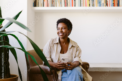 Thoughtful woman smiling on sofa, holding mobile phone and looking away