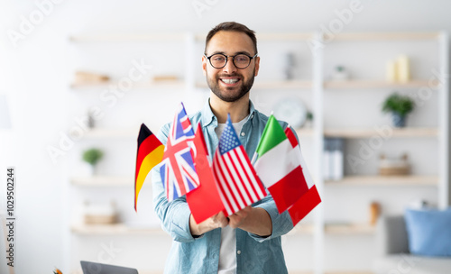 Modern education and student exchange. Cheerful young Caucasian guy holding bunch of different flags at home. Happy millennial man advertising foreign languages school, emigrating overseas