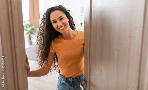 Portrait of cheerful young lady standing in doorway of new modern home, receiving and greeting visitor, happy smiling curly lady holding door looking out of slightly open ajar door