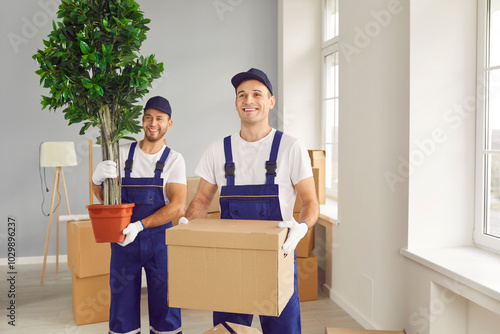 Two young smiling movers with plant and box in moving company client's new house. Caucasian male workers of moving service, dressed in work overalls, carry client's belongings into new apartment.