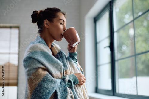 Young woman wrapped in blanket enjoying in cup of warm tea at home.