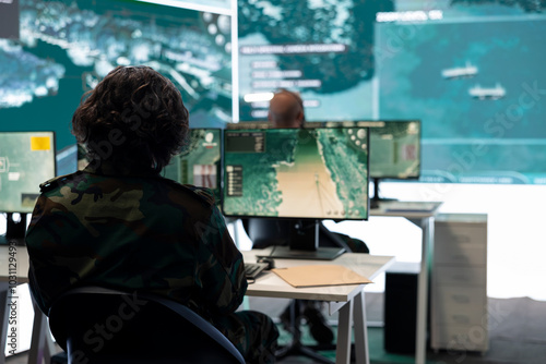 IT operator monitoring surveillance radar footage from CCTV system, using a big screen in military control room. Army recruit collecting information for missions and defense operations.