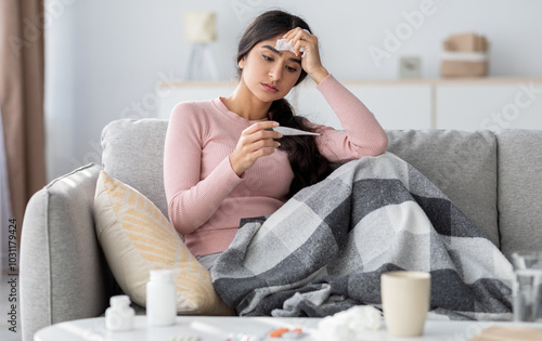 Despaired sad young hindu lady with plaid, suffer from flu, sits on sofa and looks at thermometer in living room interior. Cold, fever, disease treatment at home and covid-19 lockdown, free space