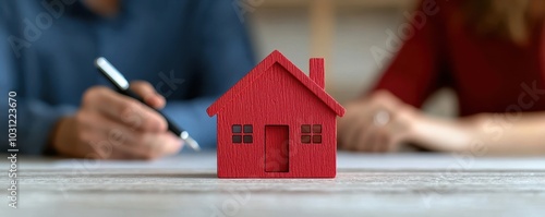 A red house model sits on a table while two people discuss paperwork, symbolizing home buying and real estate decisions.