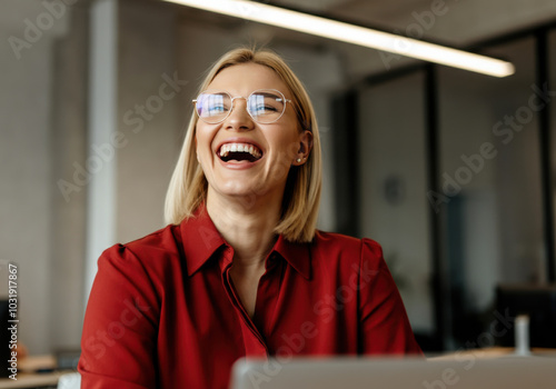 A woman in a red shirt laughs joyfully while sitting at her desk in a modern office during daytime