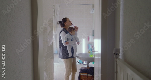 Mother multitasking in the bathroom, brushing her teeth while holding her baby. The baby gazes curiously at the surroundings, captivated by the morning routineMother multitasking in the bathroom, brus