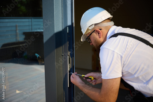 Construction worker installing door in a new house construction site. Man in working attire install front door. Builder in uniform with screwdriver adjusting front glass door in new beautiful house.