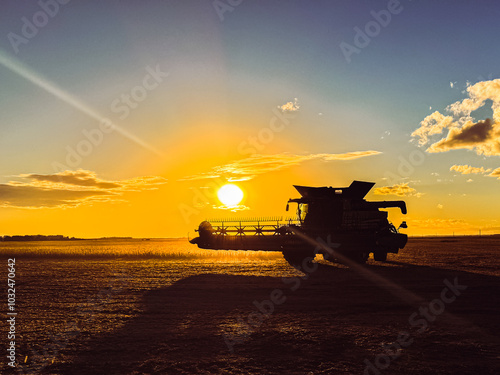 Sunset over a rural North Dakota soybean field with a combine harvester silhouetted against the sky. Sun rays shine behind the combine, highlighting the beauty of fall farming and agriculture