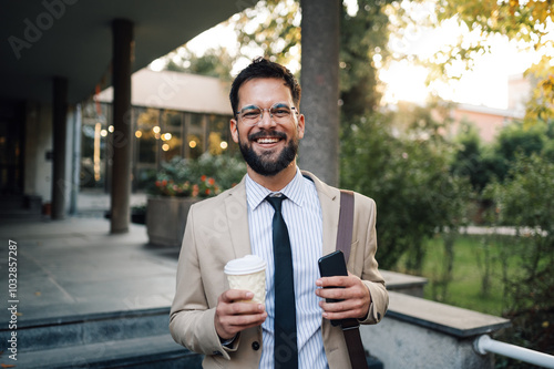 Happy businessman holding coffee and smartphone walking in the city