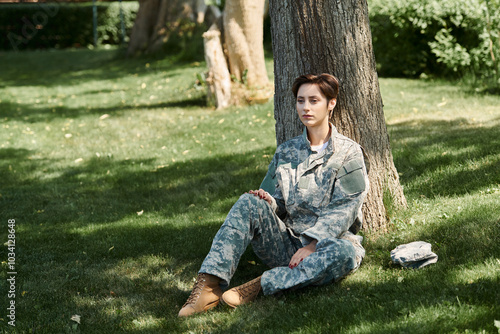 A young soldier in camouflage rests against a tree, reflecting on home after returning from service.