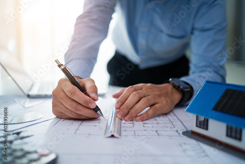 A man is drawing a house on a piece of paper with a pen. He is using a ruler to make sure the lines are straight. Concept of precision and attention to detail, as the man carefully draws the house