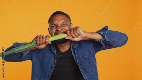 Young person biting on a green onion and acting silly in studio, pretending to eat a natural ethically sourced leek to advocate for sustainable lifestyle and veganism. Camera A.