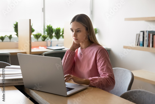 Serous thoughtful young university student girl typing on laptop in public library, thinking on report, article, essay, research paper, studying on Internet, sitting at table with open books