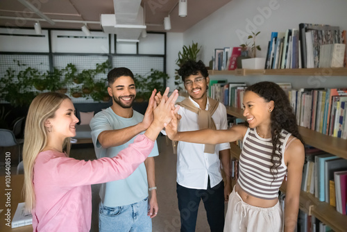 Diverse group of young students, college friends, classmates giving high five in university library space, standing at bookshelves, clapping hands in gesture of teamwork success, achievement