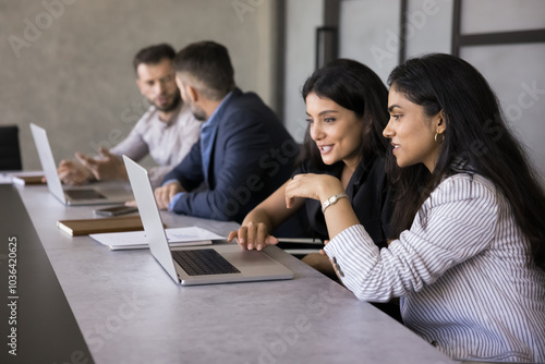 Two positive diverse Indian and Arab colleagues talking at laptop, discussing online project, watching content on Internet, speaking, smiling, meeting at large co-working workplace table for teamwork