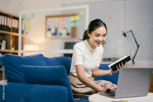 Smiling young asian businesswoman sitting on a sofa in her home office, working remotely on a laptop computer and taking notes